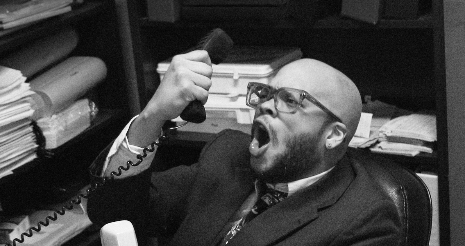 Black and white photograph of a man in a suit shouting into a telephone in a cluttered office, with a lime green jagged edge along the top.